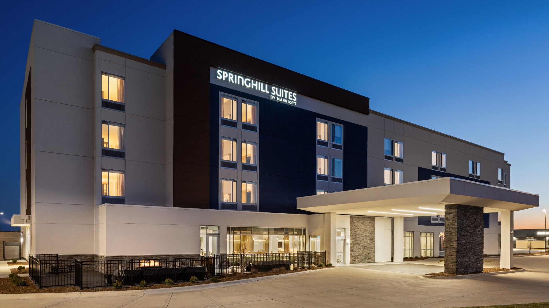Exterior of the SpringHill Suites by Marriott in Bettendorf at dusk, with a lit sign, covered entrance, and patio fire pit.