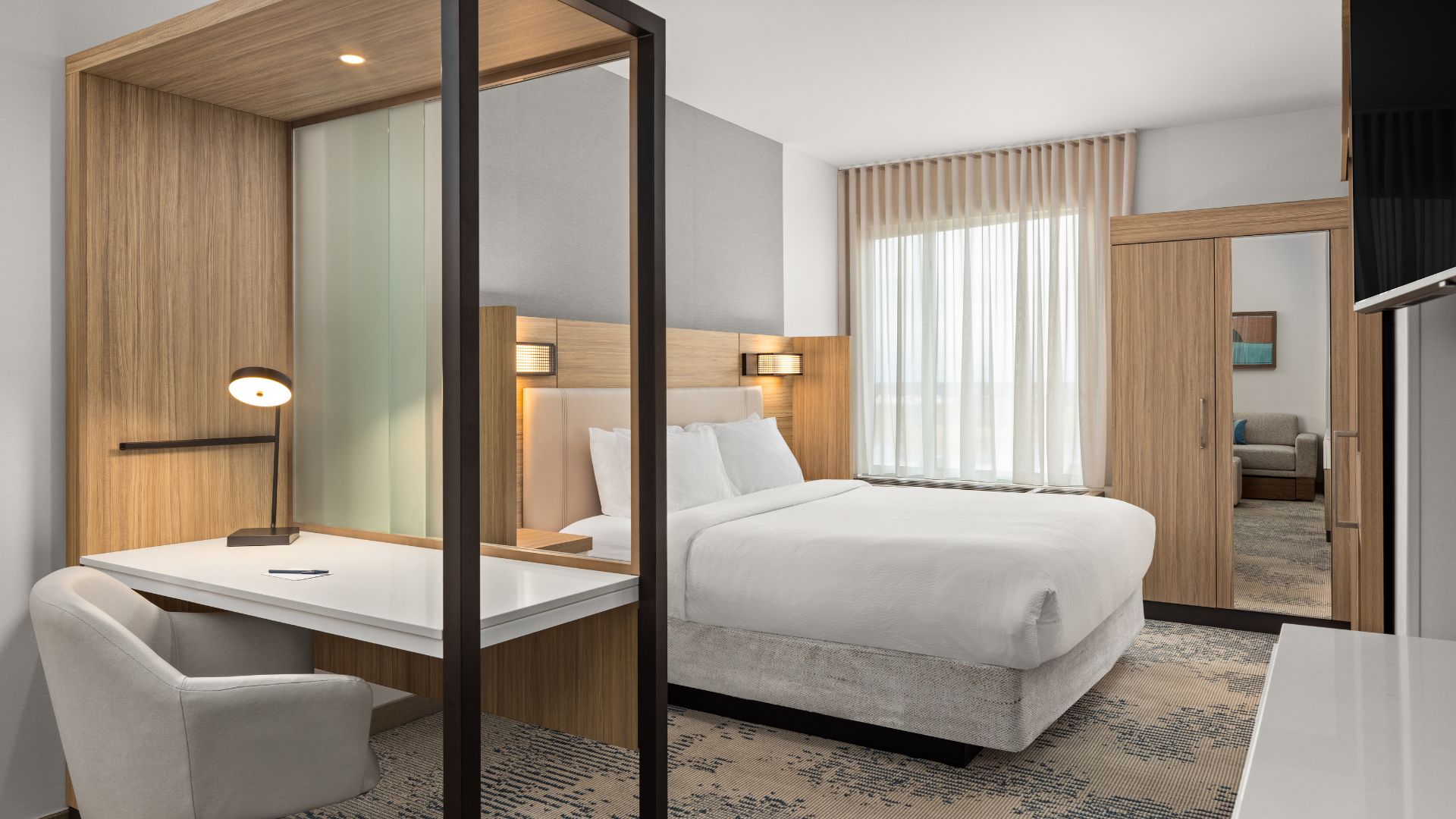Guest suite at the SpringHill Suites by Marriott in Bettendorf, with a bed, wood-paneled work desk, and sitting area beyond.