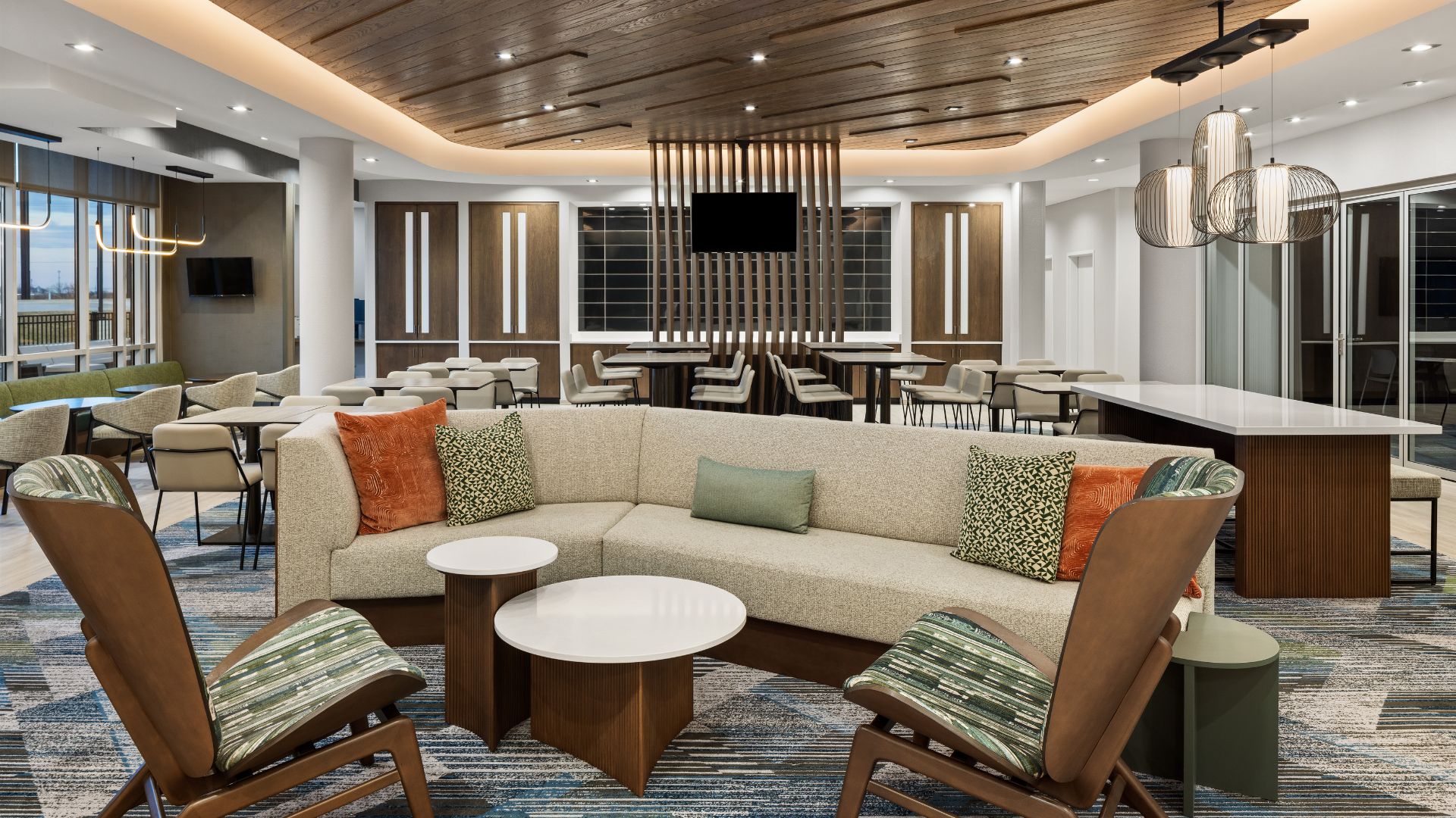 Lobby lounge at the SpringHill Suites by Marriott in Bettendorf, with a curved sofa, wood ceiling, and dining tables.