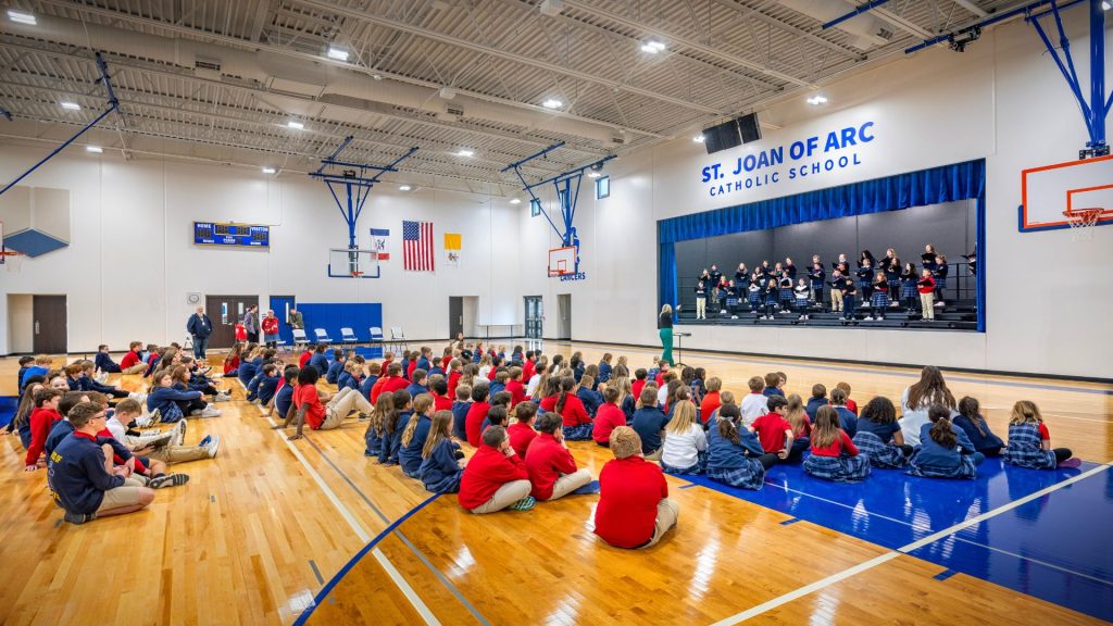 Students seated on the gym floor watching a choir perform on stage in the St. Joan of Arc Catholic School gymnasium.
