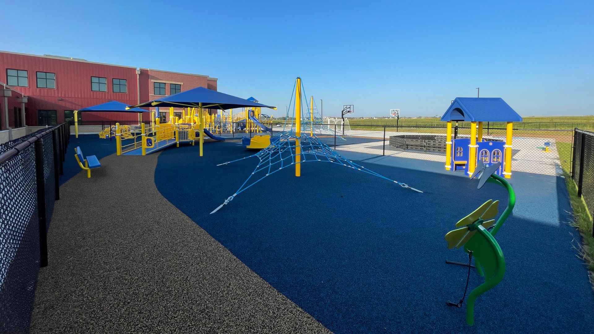 Playground at St. Joan of Arc Catholic School with a rope climbing net, shade canopies, play structures, and basketball hoops.