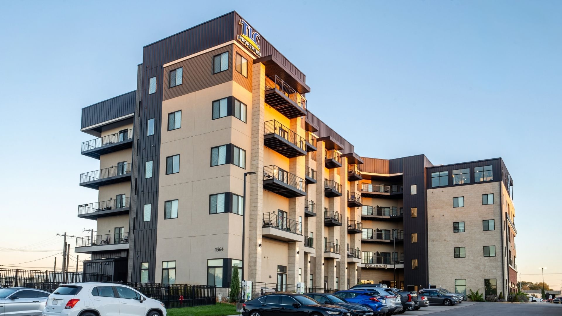 Side exterior of Heritage Flats in Springfield, Missouri, showing stacked balconies glowing at dusk above the resident parking lot