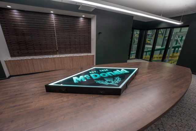 Conference room with illuminated McDonald logo inset in wood table and floor-to-ceiling windows overlooking foundry equipment.