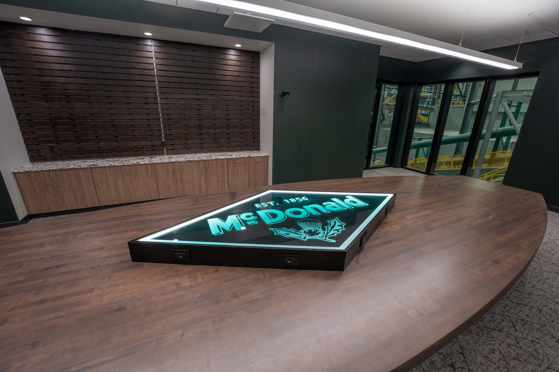 Conference room with illuminated McDonald logo inset in wood table and floor-to-ceiling windows overlooking foundry equipment.
