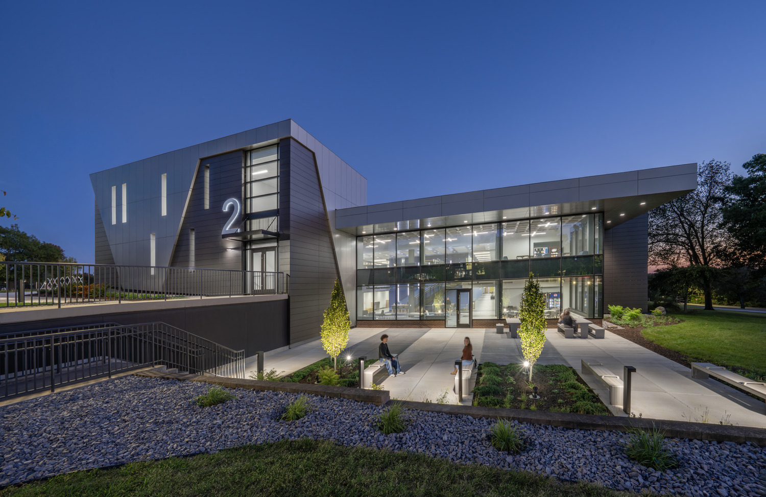 Black Hawk College Building 2 at dusk with an illuminated glass entry, outdoor seating, and landscaped courtyard