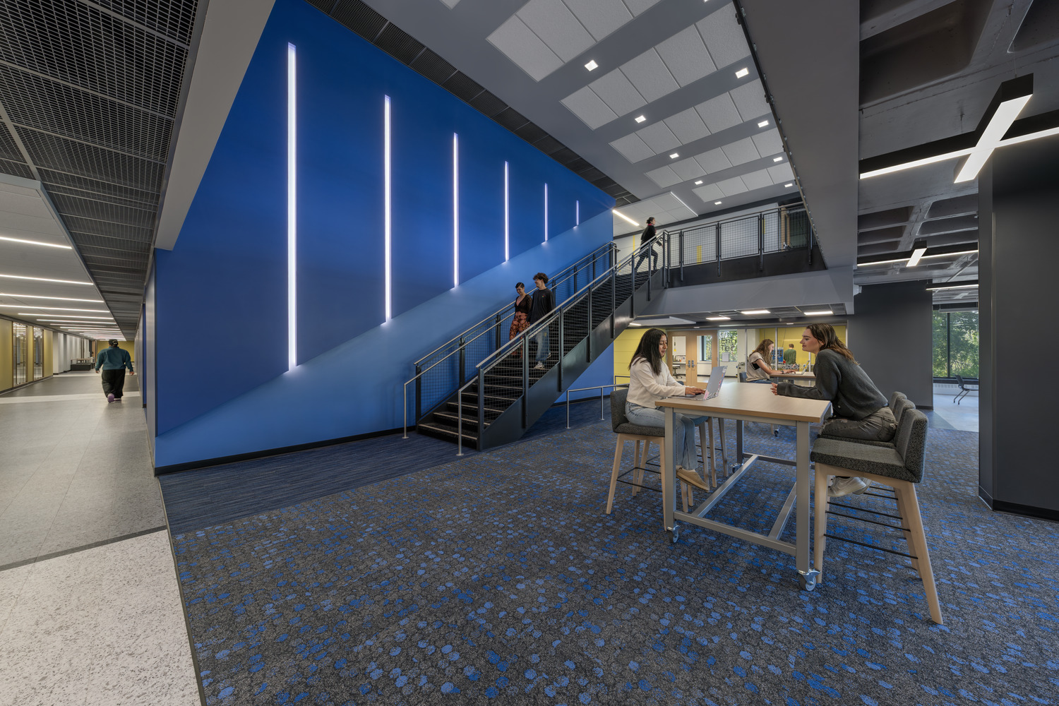 Students study at a high-top table near a blue feature wall and open staircase in Black Hawk College Building 2